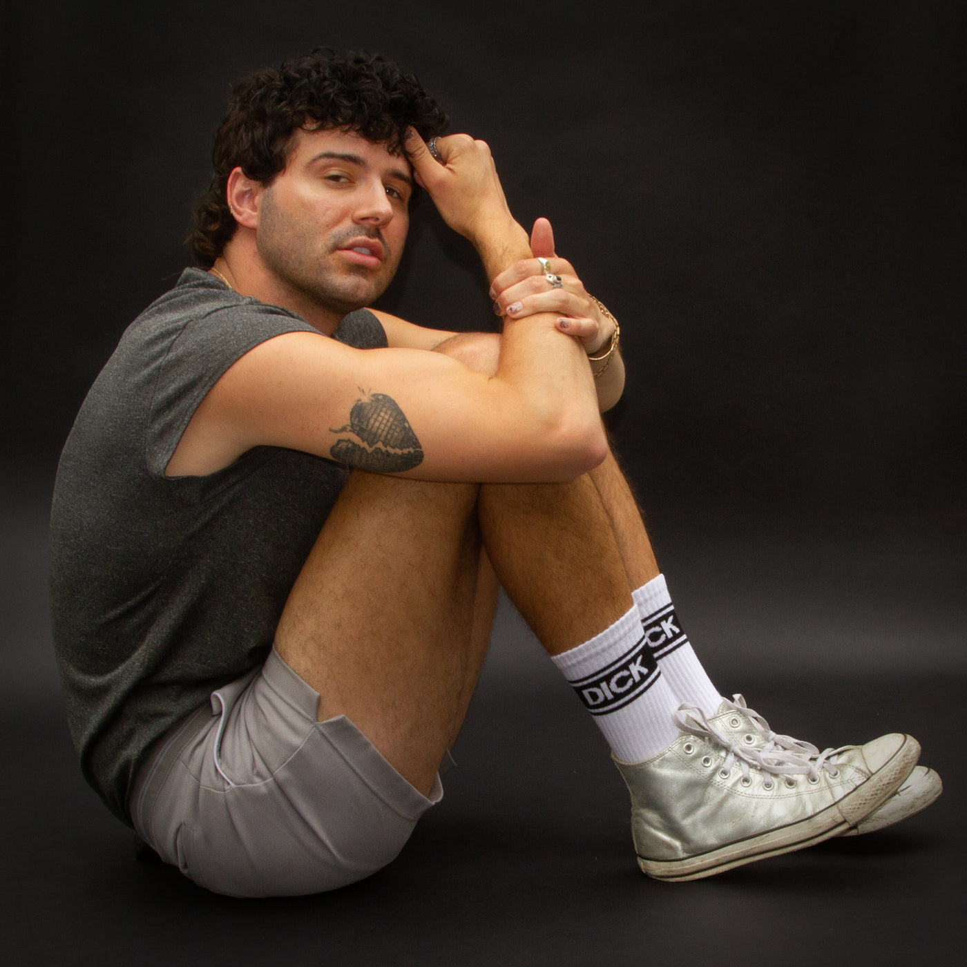 Lifestyle shot: A person sitting with their knees up, wearing 'Dick' socks and silver Converse sneakers. [lf]