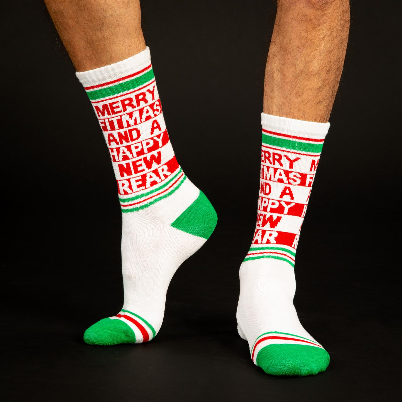 Lifestyle shot of a person's legs wearing the "Merry Fitmas and a Happy New Rear" socks against a black background. [lf]