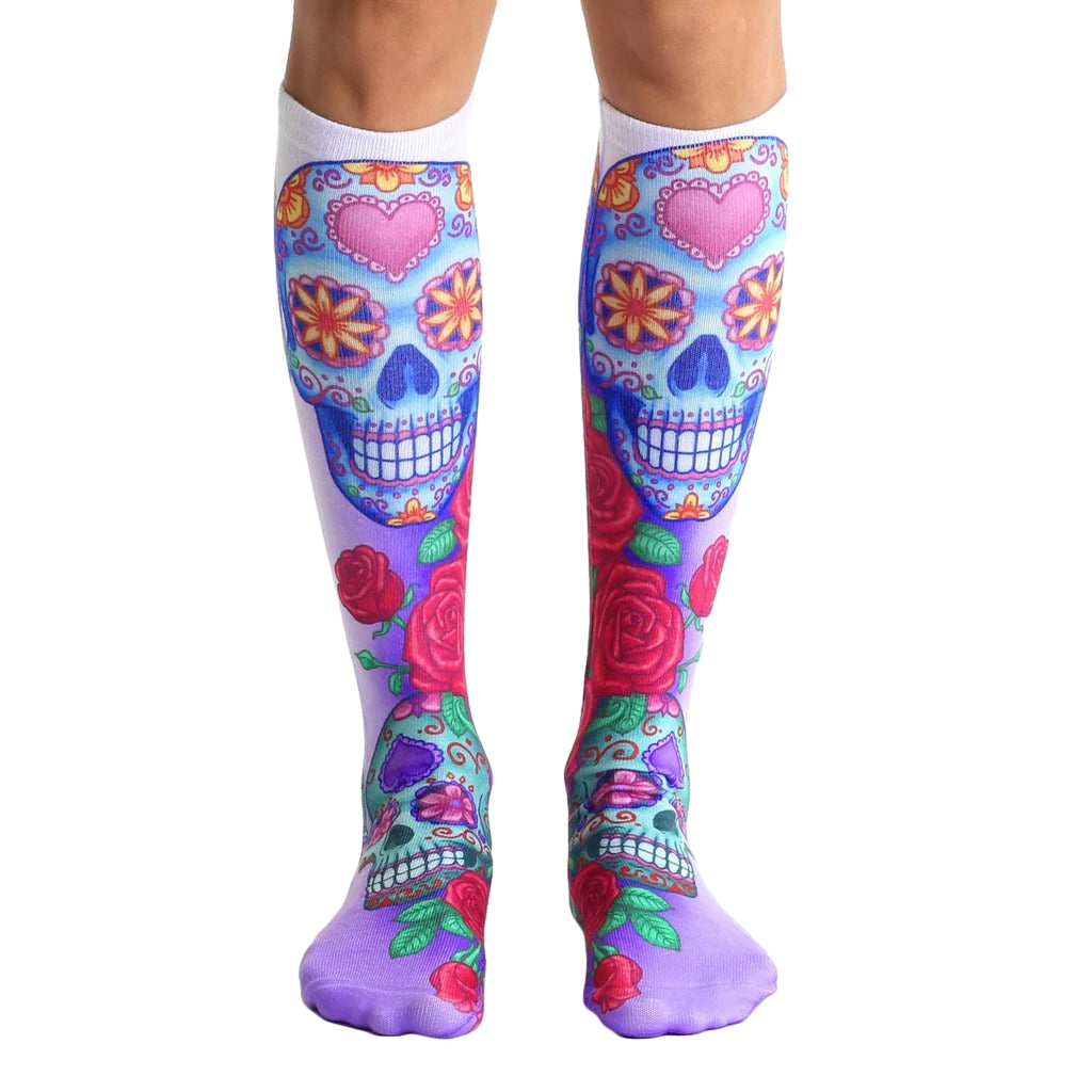 Pair of knee-high socks.  The main design features colorful sugar skulls with pink hearts and roses on a white and purple background. [lf]